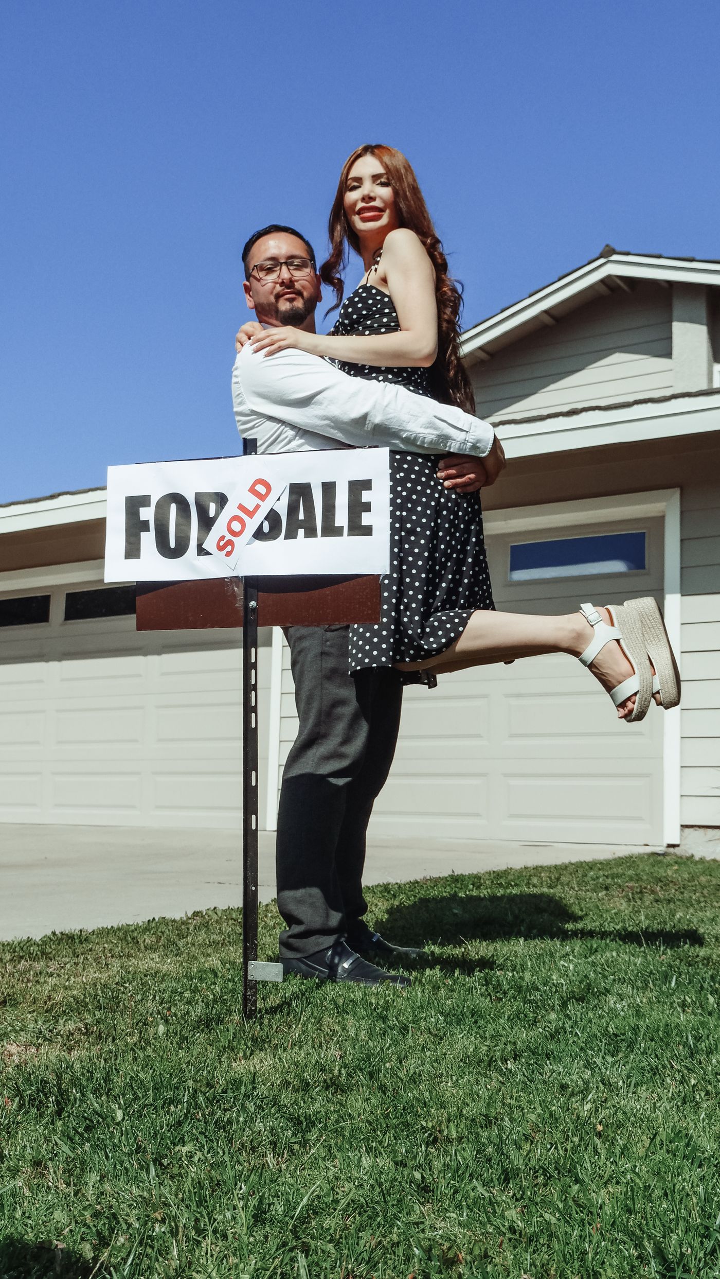 A happy couple in front of their new home by a For Sale sign marked Sold